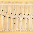 A close-up of a cream-colored fringe curtain or wall hanging with embroidered olive branches and small brown beads, shown against a warm yellow background.