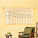 A cream macrame wall hanging with embroidered green leaves and small wooden beads, displayed on a wooden rod on a yellow wall above a green armchair.