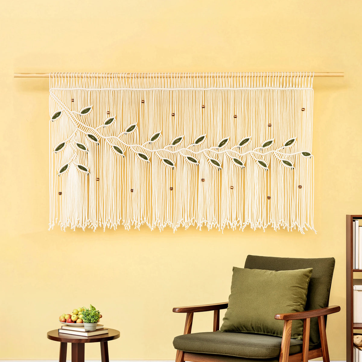 A cream macrame wall hanging with embroidered green leaves and small wooden beads, displayed on a wooden rod on a yellow wall above a green armchair.