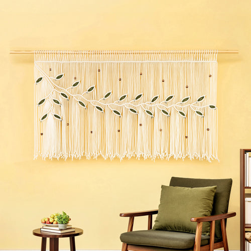 A cream macrame wall hanging with embroidered green leaves and small wooden beads, displayed on a wooden rod on a yellow wall above a green armchair.
