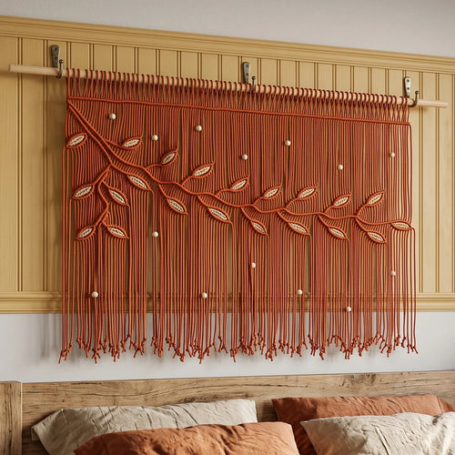 A decorative window curtain panel with hanging string and embroidered leaf motifs, shown installed above a bed in a warm beige bedroom.