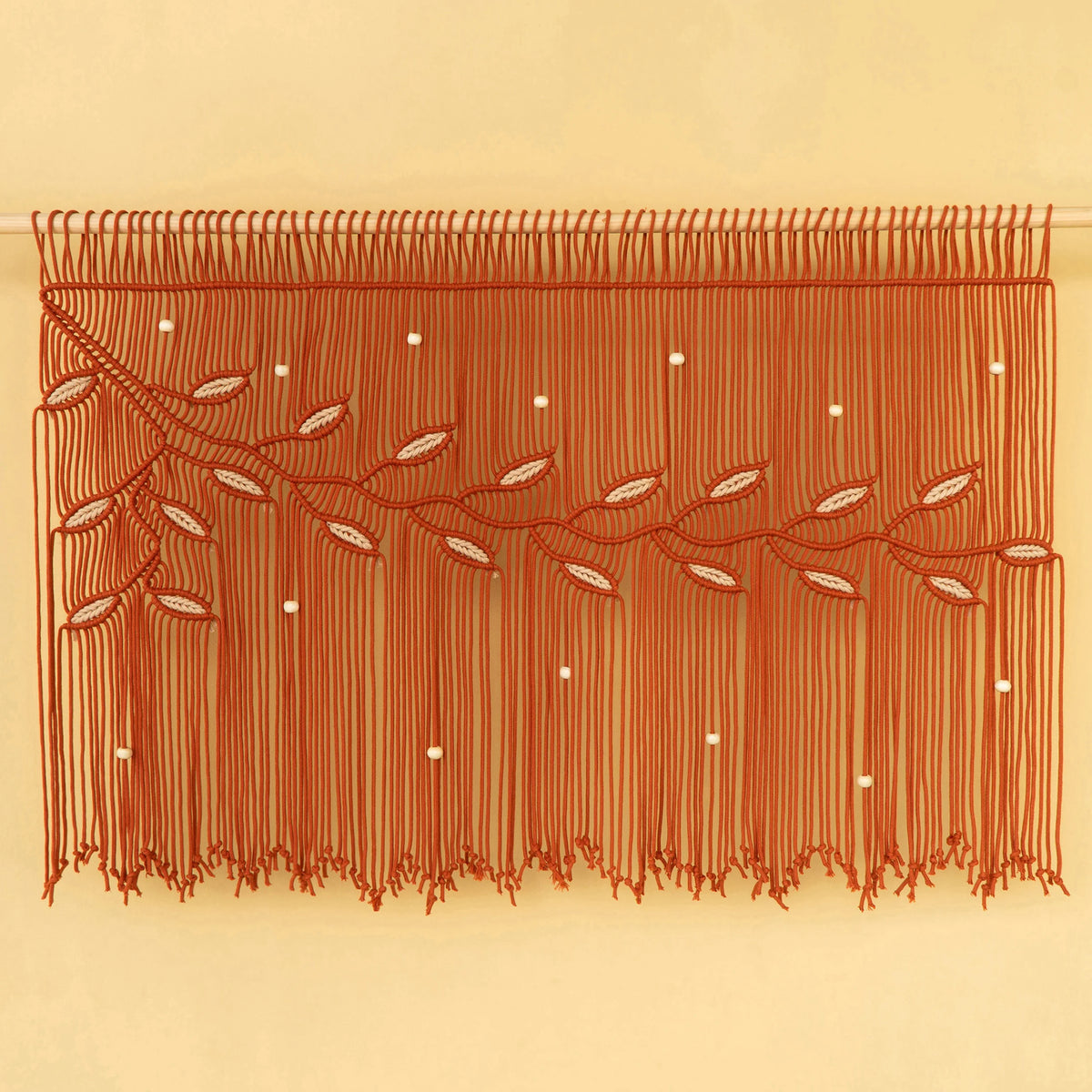 A rust-orange macrame wall hanging with woven branches, leaf patterns, and small white bead accents, suspended against a solid beige background.