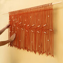 A terracotta orange macrame wall hanging with leaf and bead details, suspended from a light-colored wooden rod on a beige wall, with hands adjusting it.