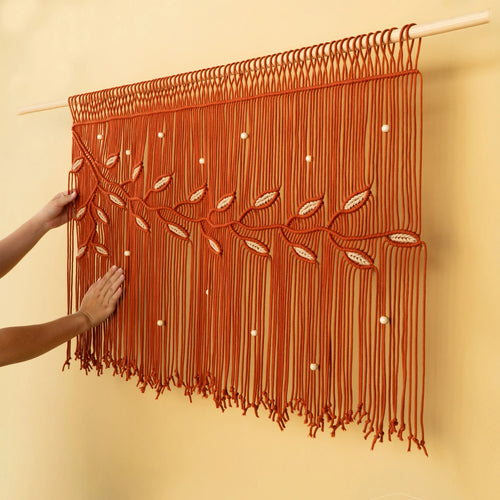 A terracotta orange macrame wall hanging with leaf and bead details, suspended from a light-colored wooden rod on a beige wall, with hands adjusting it.