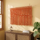 A bohemian-style woven wall hanging with green fringes, embroidered leaf motifs, and hanging beads, displayed on a beige wall above a wooden console table.