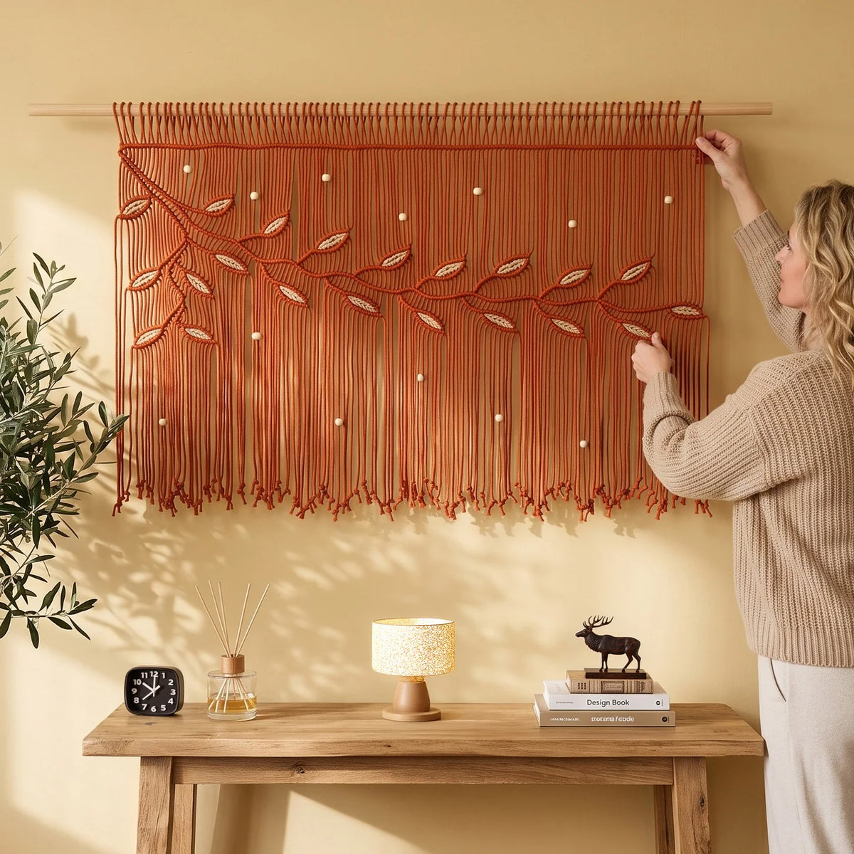 A woman hangs a large woven macrame wall hanging with leaf and vine motifs on a yellow wall above a wooden console table. The room has warm sunlight, a potted tree, and umbrellas in the background.