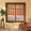 A rust-orange macrame valance with a leafy branch pattern hanging in a dark wood window frame, styled in a warm, modern living room setting with a potted plant and an orange armchair.