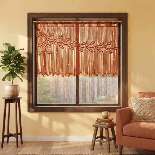 A rust-orange macrame valance with a leafy branch pattern hanging in a dark wood window frame, styled in a warm, modern living room setting with a potted plant and an orange armchair.