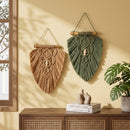 Two bohemian macrame wall hangings, one in tan and one in sage green, displayed on a beige wall above a wooden cabinet with a plant and books.