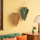 Two bohemian macrame wall hangings, one in natural tan and the other in sage green, displayed on a warm yellow wall in a styled living room setting.