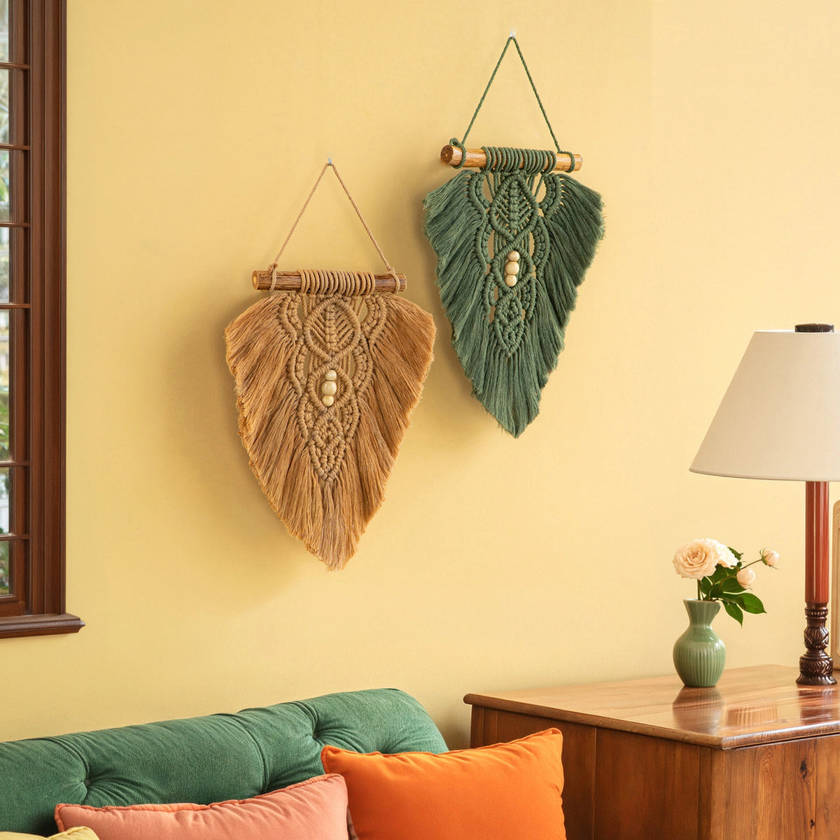 Two bohemian macrame wall hangings, one in natural tan and the other in sage green, displayed on a warm yellow wall in a styled living room setting.