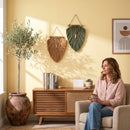 A woman sits in a beige armchair in a sunlit living room, holding a mug. Two woven wall hangings in brown and green hang on a pale yellow wall above a wooden sideboard with books and a small plant.