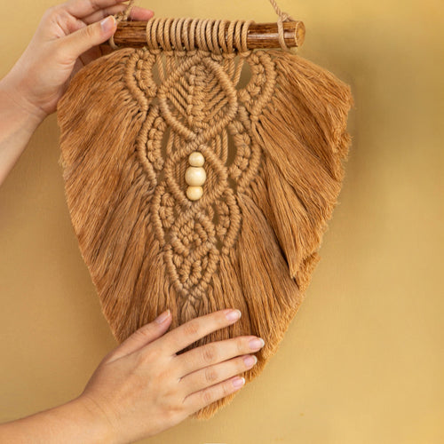 A hand-woven bohemian macrame wall hanging in a warm tan color, held up by two hands against a solid tan background. The hanging features intricate woven patterns, a wooden dowel at the top, and three small wooden beads in the center.