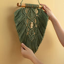 A green macrame wall hanging with a wooden dowel and tassel, held by two hands against a warm beige background.