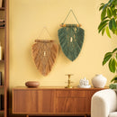 Two decorative macrame wall hangings are displayed on a warm yellow wall above a wooden console table. One hanging is natural tan with a central white bead, and the other is dark green with a central white bead.