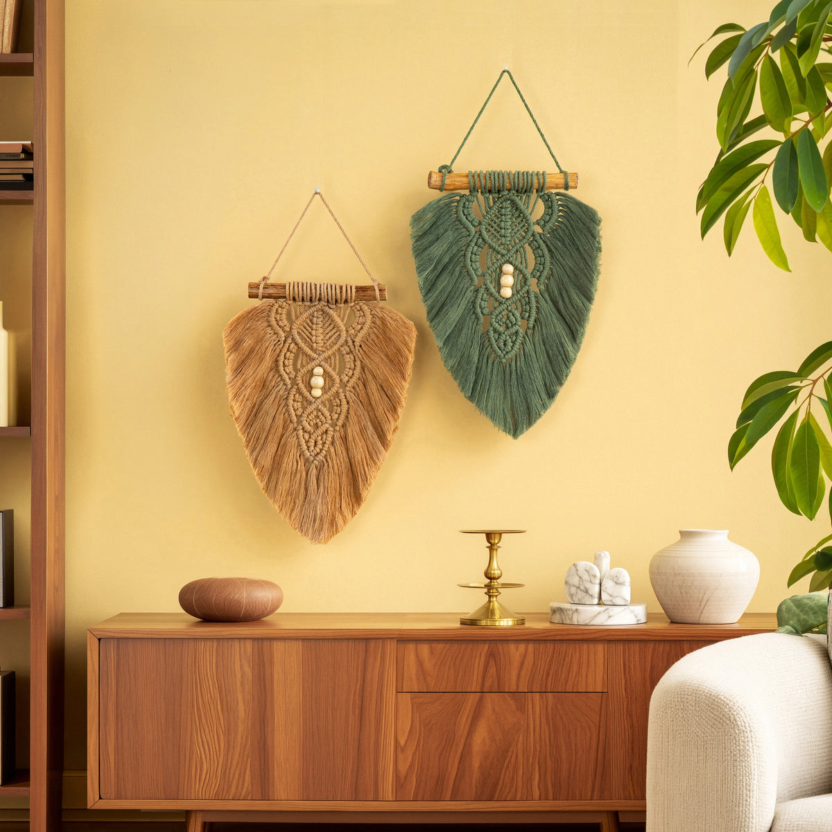 Two decorative macrame wall hangings are displayed on a warm yellow wall above a wooden console table. One hanging is natural tan with a central white bead, and the other is dark green with a central white bead.