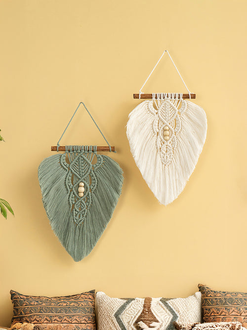 Two macrame wall hangings on a yellow wall with patterned pillows below.