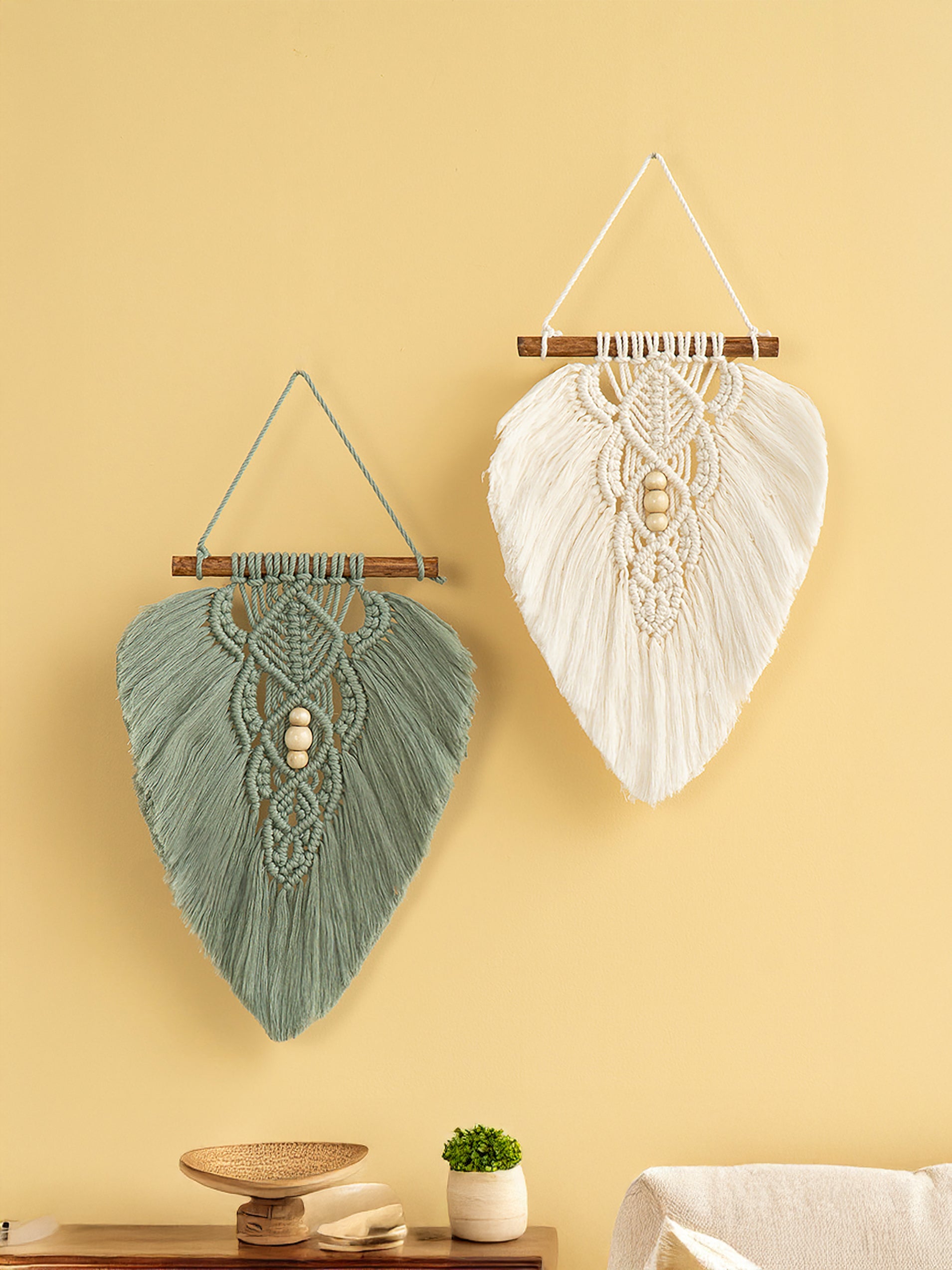 Two macrame wall hangings on a yellow wall with a plant and decorative object below.