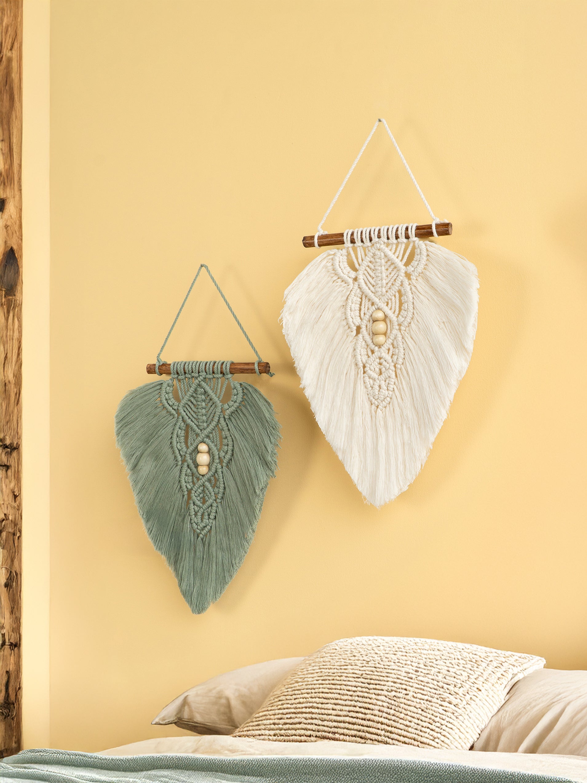 Two macrame wall hangings, one green and one white, on a yellow wall.