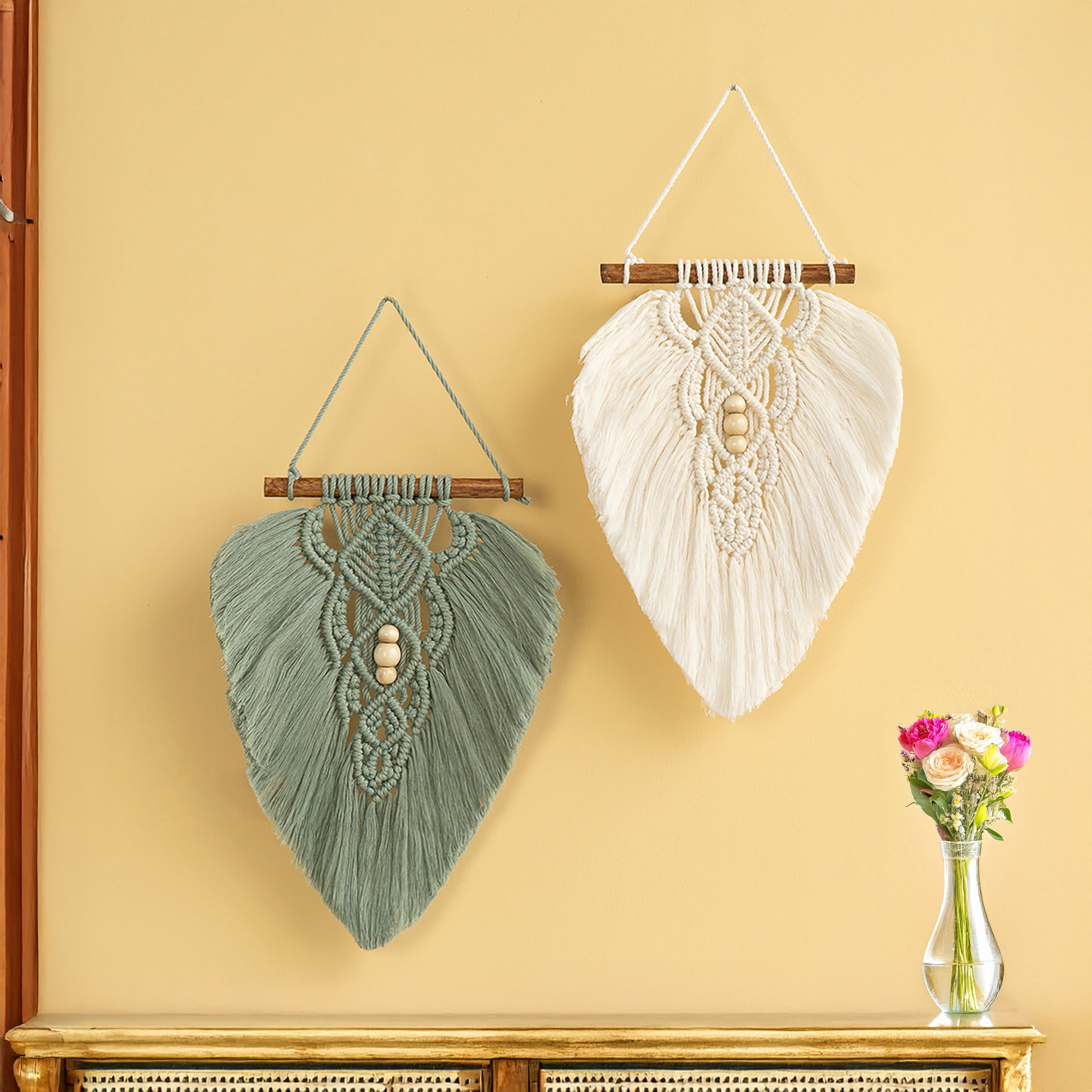 Two macrame wall hangings on a yellow wall with a vase of flowers below.