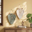 Two bohemian macrame wall hangings, one sage green and one cream, displayed on a warm yellow wall above a wooden console table with a plant and books.