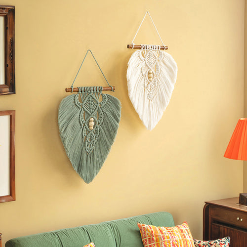 Two leaf-shaped macrame wall hangings, one sage green and one white, displayed on a warm beige wall above a green sofa with patterned pillows.