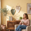 A woman sits in a beige armchair in a warm, sunlit room, holding a ceramic mug. Two leaf-shaped macrame wall hangings are displayed on a yellow wall above a wooden cabinet, next to a potted olive tree.