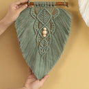 A sage green macrame wall hanging with intricate woven patterns, hanging tassels, and three wooden beads at the center, held up by hands against a warm beige background.