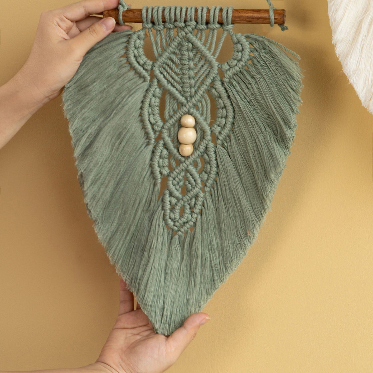 A sage green macrame wall hanging with intricate woven patterns, hanging tassels, and three wooden beads at the center, held up by hands against a warm beige background.