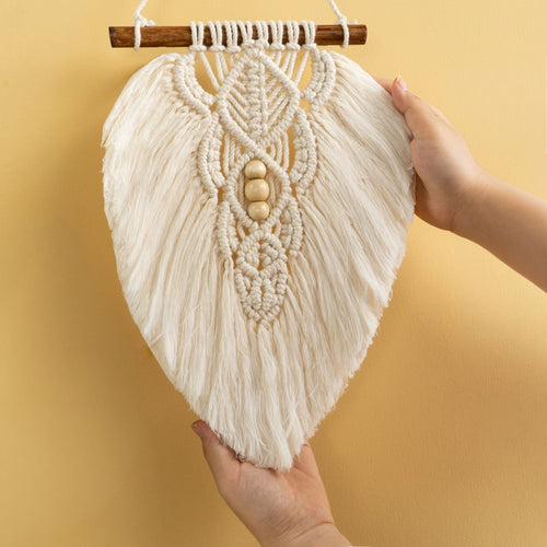 A large off-white macrame wall hanging with intricate woven patterns, wooden beads, and a wooden dowel, held up by two hands against a warm yellow background.