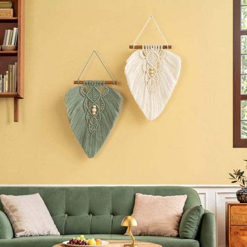 Two leaf-shaped macrame wall hangings, one sage green and one cream, displayed on a yellow wall above a green sofa in a styled living room.