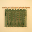 A green macrame wall hanging with leafy vine patterns and tassel fringe, suspended from a wooden rod against a warm beige wall.