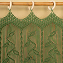 Green macrame wall hanging with a leaf pattern, suspended from a wooden rod with white rings against a beige background.