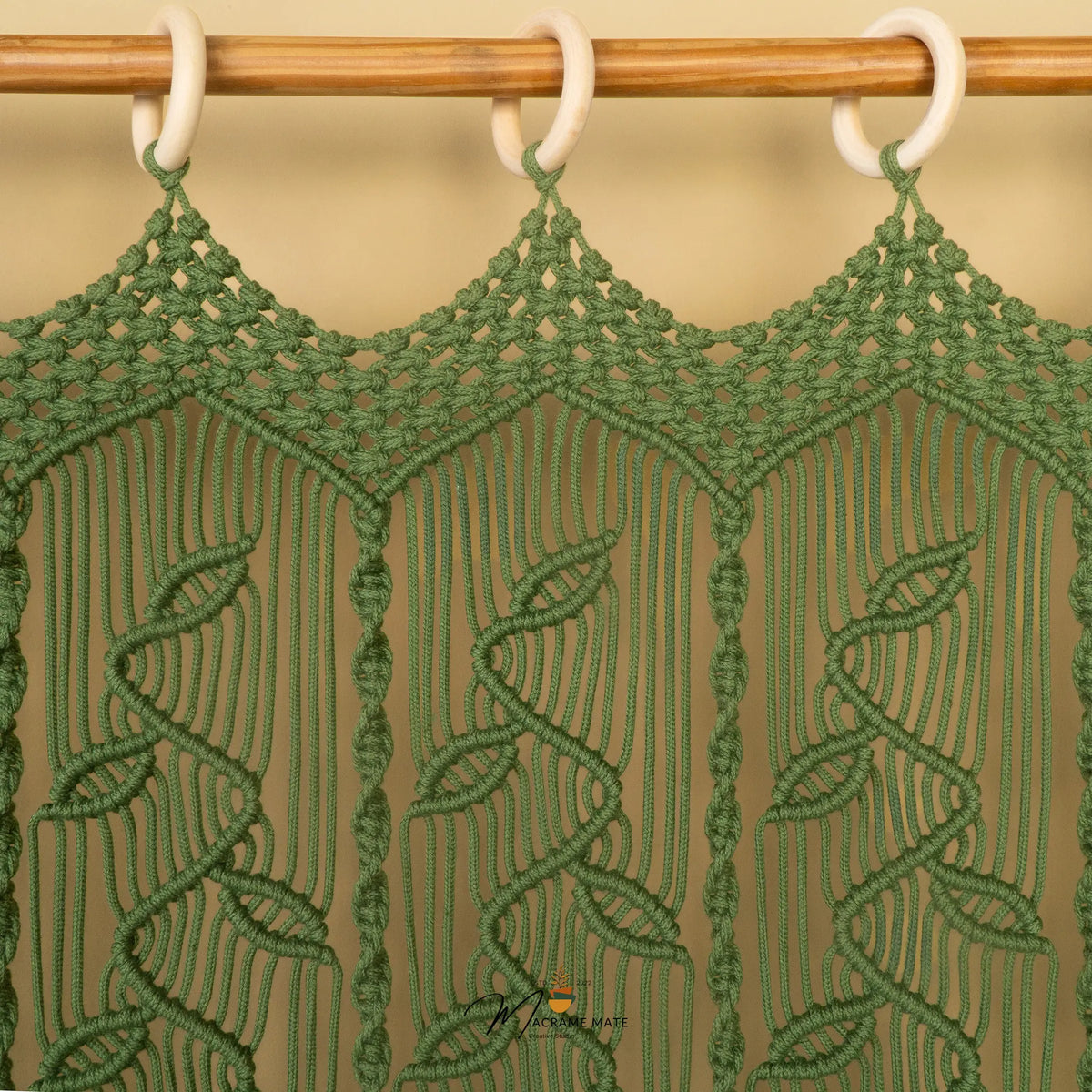 Green macrame wall hanging with a leaf pattern, suspended from a wooden rod with white rings against a beige background.
