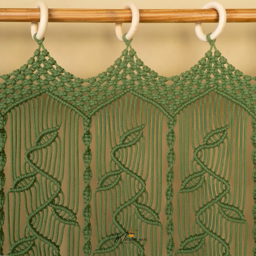 Green macrame wall hanging with a leaf pattern, suspended from a wooden rod with white rings against a beige background.