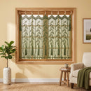 A set of three window curtains with a green leaf pattern on a cream background, hanging in a wooden window frame in a styled room with a potted plant and an armchair.