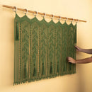 An olive green macrame wall hanging with intricate woven patterns and tassels, suspended from a wooden dowel with white rings on a beige wall. Two hands are shown adjusting the hanging on the right side.