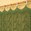 A green macrame curtain panel hanging from a wooden rod with white plastic rings against a warm beige wall.