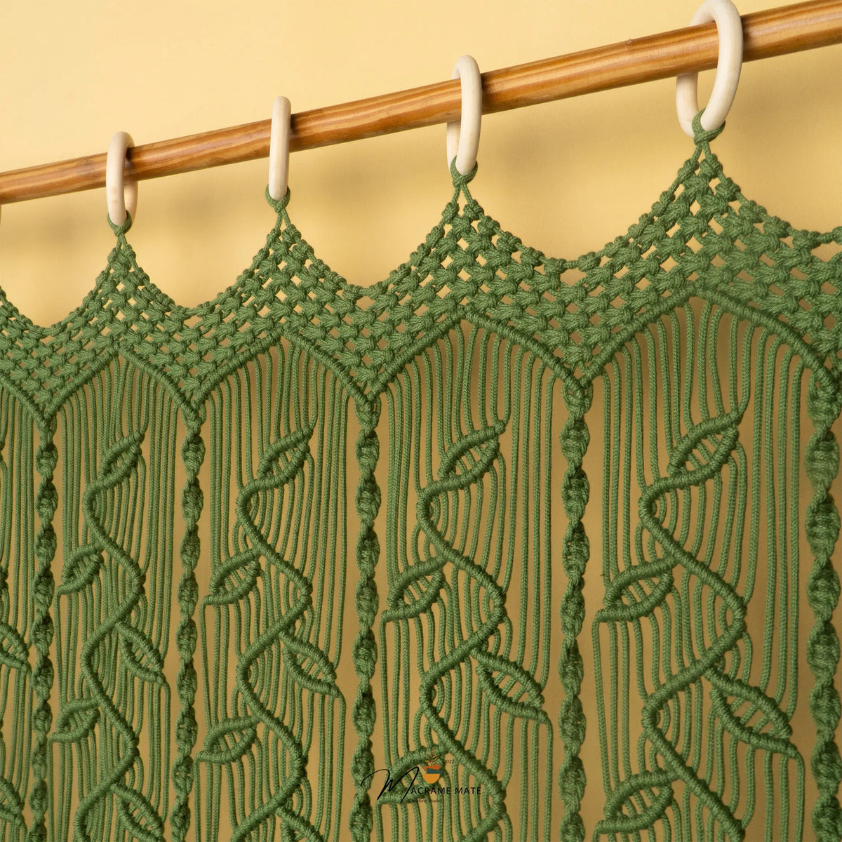 A green macrame curtain panel hanging from a wooden rod with white plastic rings against a warm beige wall.