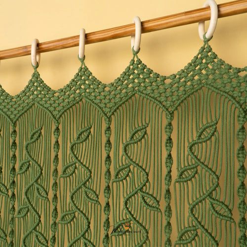 A green macrame curtain panel hanging from a wooden rod with white plastic rings against a warm beige wall.