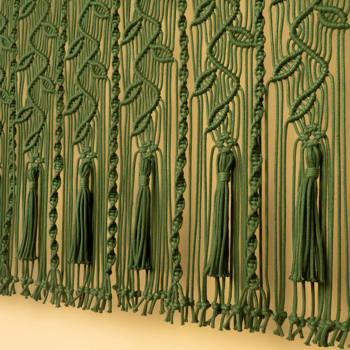 Green macrame wall hanging with knotted leaf patterns and tassels, shown against a beige wall.