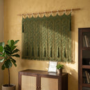An olive green macrame wall hanging with intricate woven patterns and tassels, suspended from a wooden rod on a textured yellow wall in a styled room setting.
