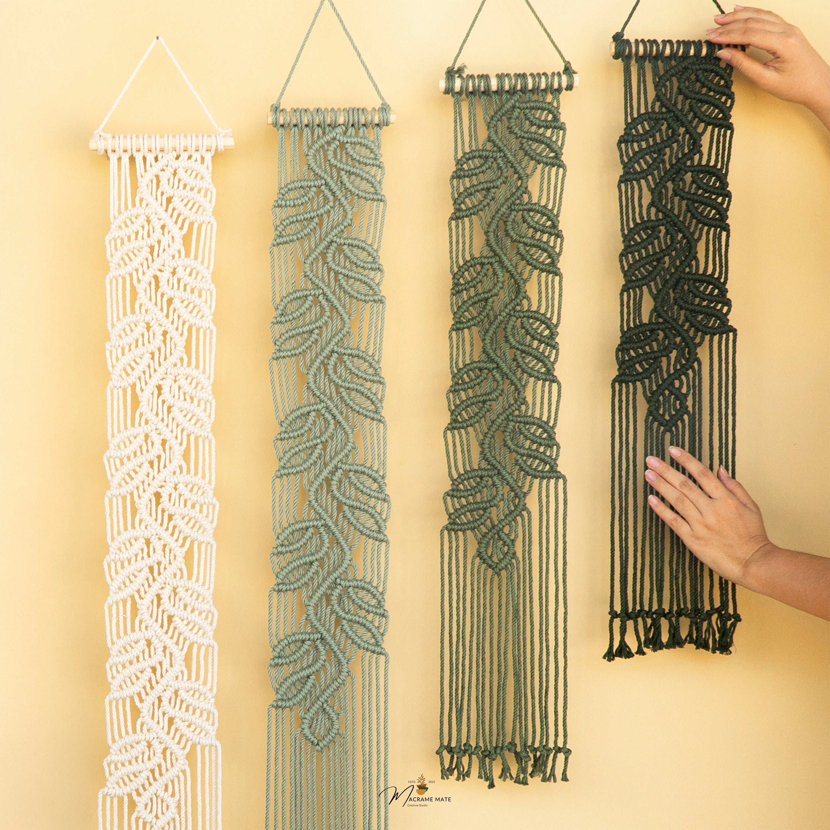 Four macrame wall hangings with leaf patterns in white, sage green, olive green, and black, displayed against a warm beige background. Two hands are shown holding the black and green hangings to demonstrate scale and texture.