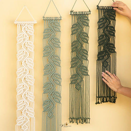 Four macrame wall hangings with leaf patterns in white, sage green, olive green, and black, displayed against a warm beige background. Two hands are shown holding the black and green hangings to demonstrate scale and texture.