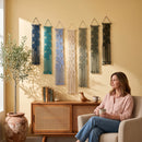A woman sits in a beige armchair holding a mug in a sunlit room, with six woven wall hangings in assorted colors displayed above a wooden cabinet. A potted olive tree stands on the left.