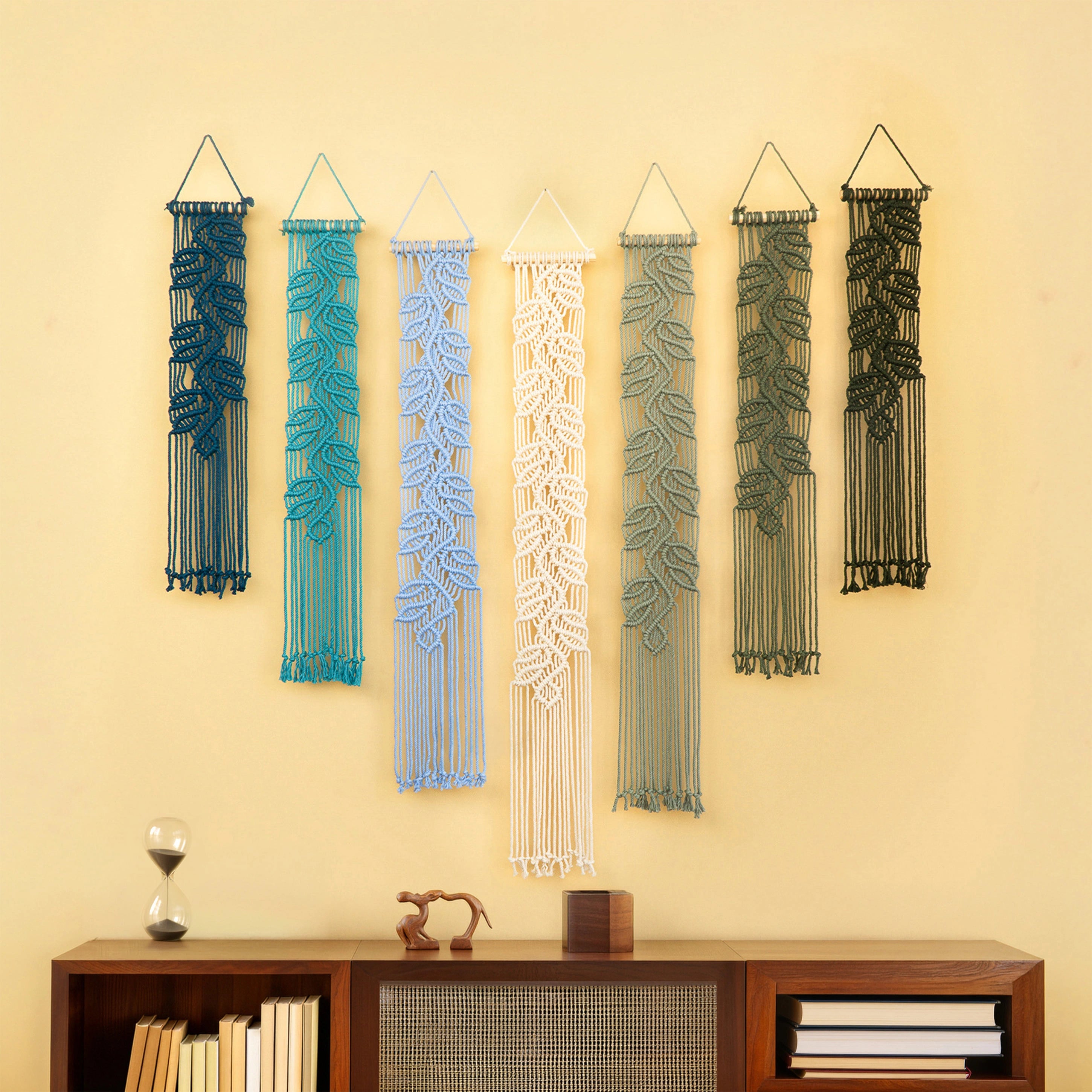 Six macrame wall hangings with leaf patterns in dark blue, teal, light blue, white, sage green, and olive green, displayed on a beige wall above a wooden console table.
