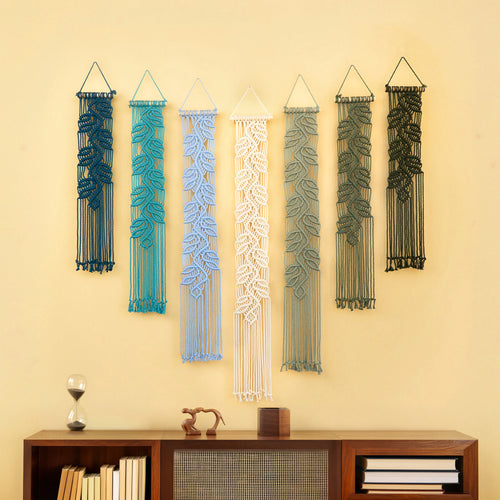 Six macrame wall hangings with leaf patterns in dark blue, teal, light blue, white, sage green, and olive green, displayed on a beige wall above a wooden console table.