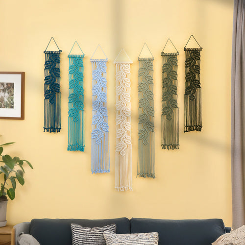 Six macrame wall hangings with leaf patterns in various colors (dark blue, teal, light blue, cream, sage green, and black) are displayed on a yellow wall above a dark blue sofa in a living room setting.