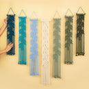 Six macrame wall hangings in different colors are displayed against a warm beige background. A person's hands are visible on the left, holding the dark blue one to show scale.