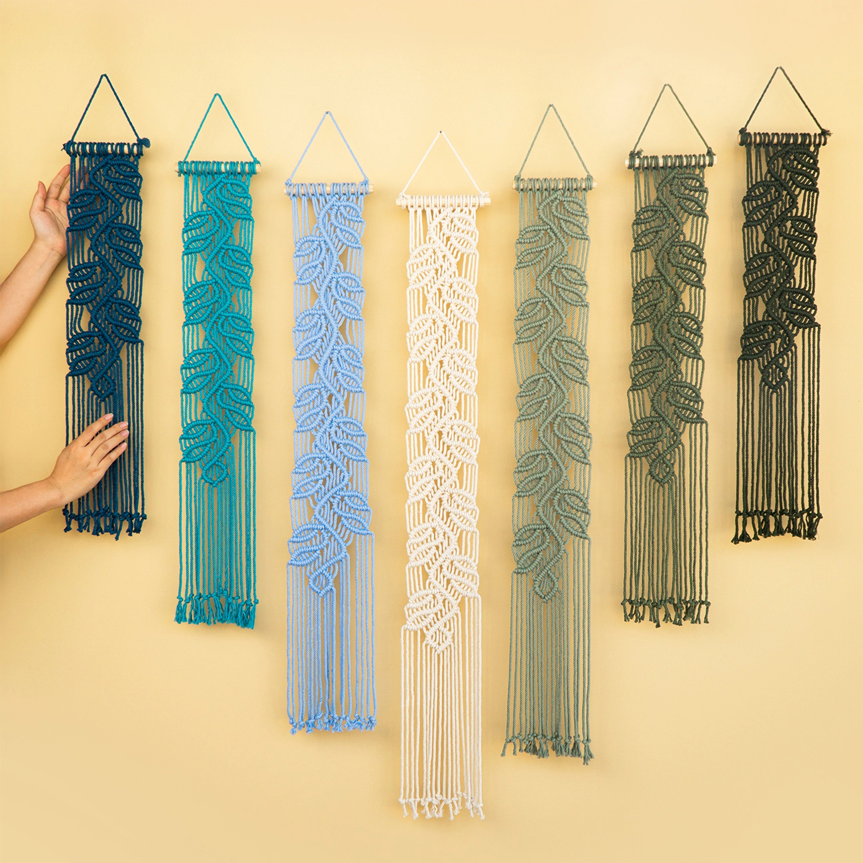 Six macrame wall hangings in different colors are displayed against a warm beige background. A person's hands are visible on the left, holding the dark blue one to show scale.